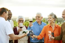 Man pouring red wine for senior males and females during sunset. Smiling elders enjoying winetasting in vineyard. They are standing against sky.