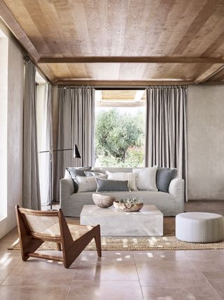 neutral living room with outside view, stone flooring, grey sofa, wooden chairs, taupe curtains wooden ceiling