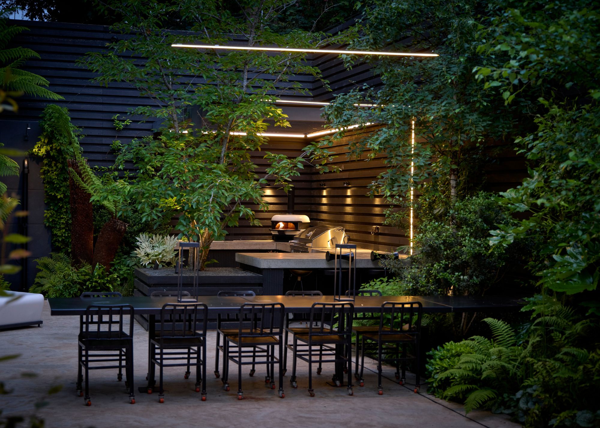 An outdoor kitchen with strip lighting around it and wall lights along the middle section with a gozney pizza oven and a long black table and chairs surrounded by green foliage