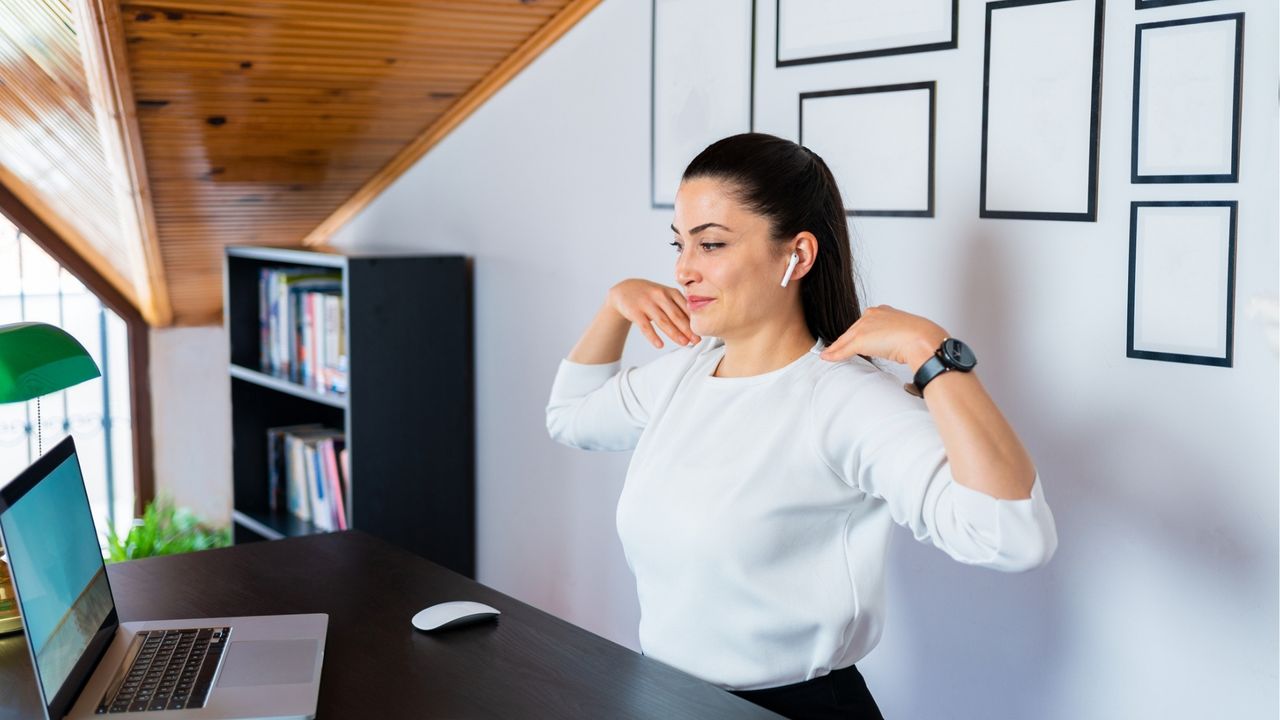 A woman in white long sleeved top with fingertips on shoulders in front of laptop with an office background