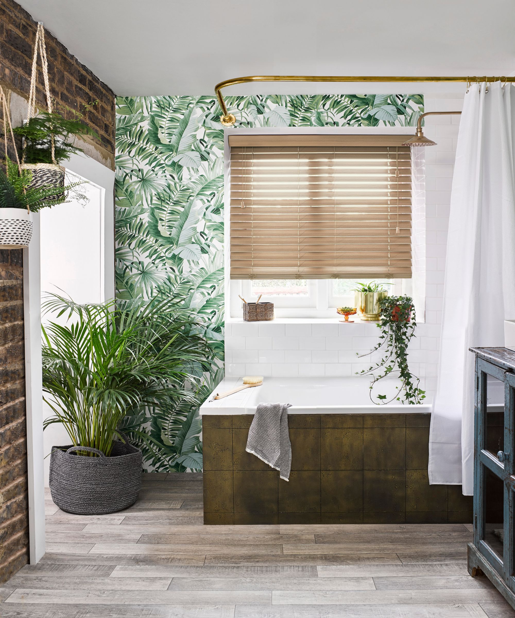 A vibrant, jungle-inspired bathroom with bold tropical leaf wallpaper and an exposed brick wall. A built-in bath with a dark olive tile panel sits beneath a brass shower rail and white curtain, surrounded by lush hanging and potted palms on wood-effect floor tiles.