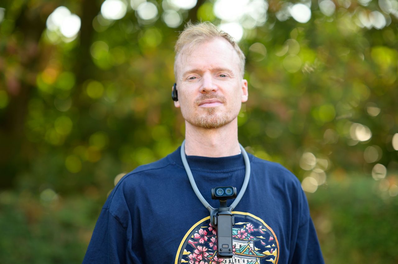 Tim Coleman wearing the Insta360 POV Head Tracker accessory in his ear, and Luna Ultra camera on his chest attached to a neck mount, outdoors with dappled light in background foliage