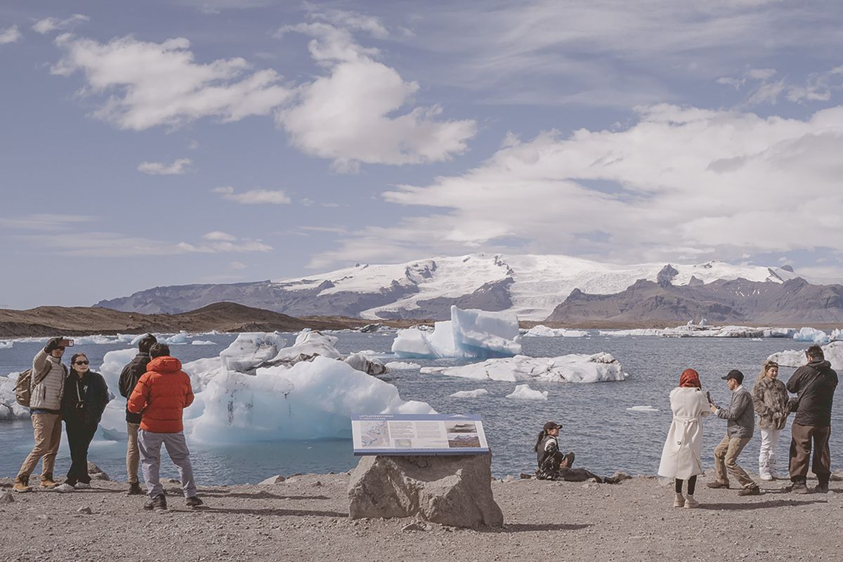 Tourists pose for selfies and photos on a gravel shore beside a lagoon full of floating icebergs, with a glacier and snow-capped mountains in the distance and a