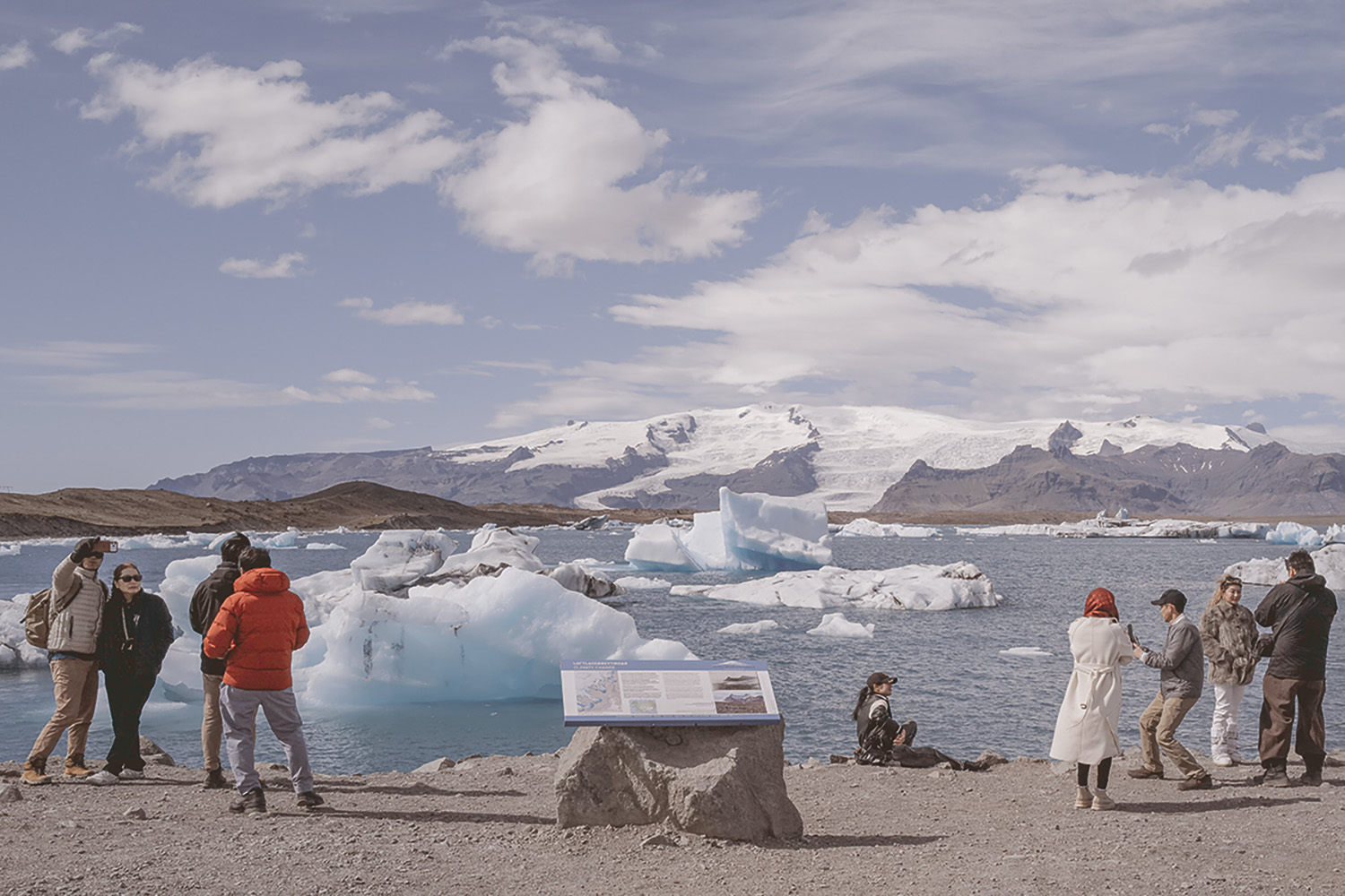 Tourists pose for selfies and photos on a gravel shore beside a lagoon full of floating icebergs, with a glacier and snow-capped mountains in the distance and an information board in the foreground.
