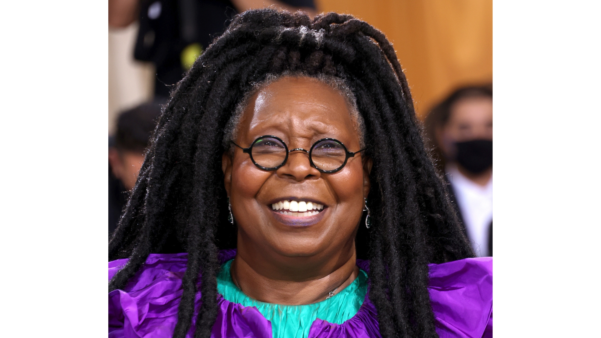 Whoopi Goldberg pictured at the 2021 Met Gala smiling, wearing a purple and turquoise dress and round glasses
