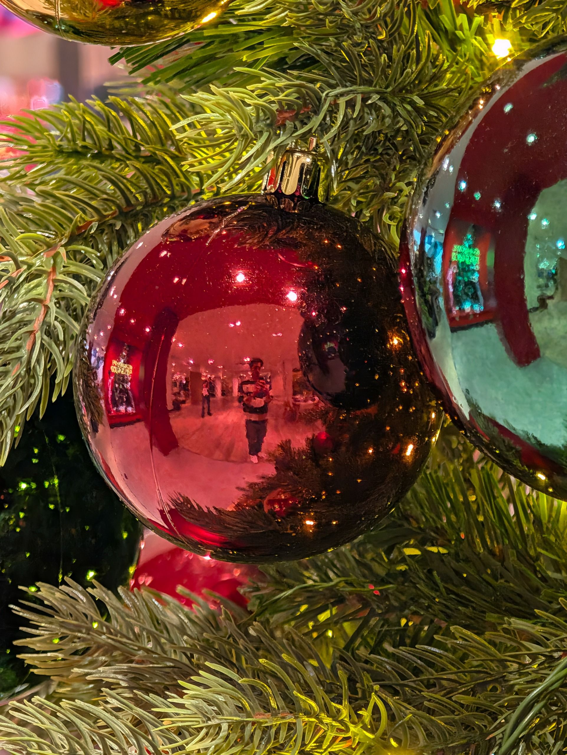Close-up of a shiny red Christmas bauble reflecting the photographer and surrounding festive decorations.