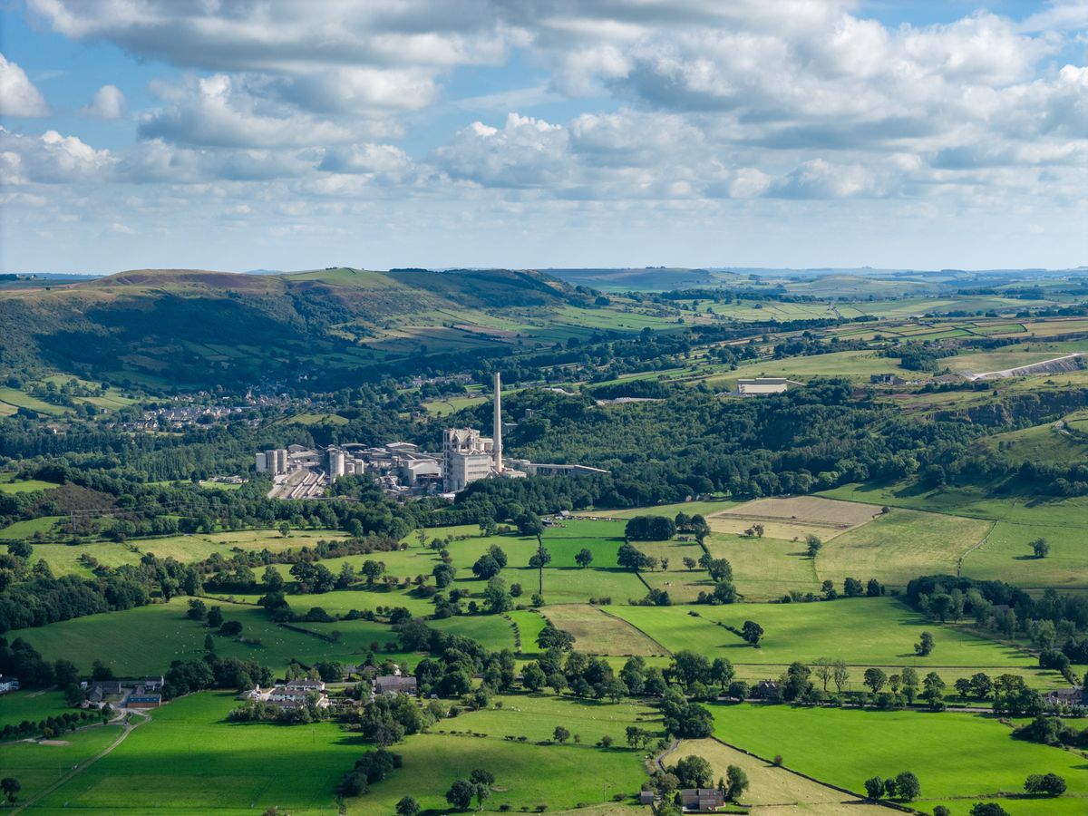 Image taken by DJI Air 3 (a camera drone). Here the focus is an industrial plant in the distance. It is surrounded by dark green trees. In the distance are rolling green hills dotted with dark green trees and a gray, cloudy sky.