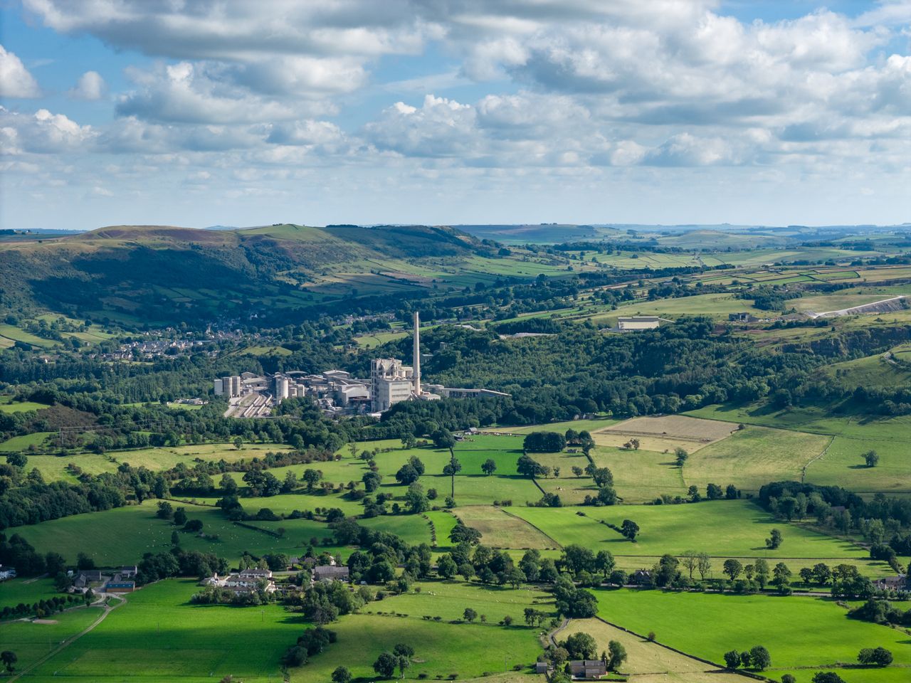 Image taken by DJI Air 3 (a camera drone). Here the focus is an industrial plant in the distance. It is surrounded by dark green trees. In the distance are rolling green hills dotted with dark green trees and a gray, cloudy sky.