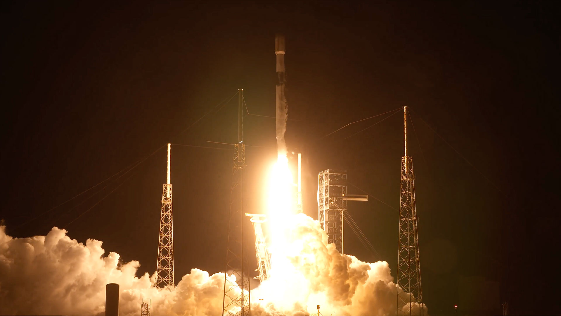 Um foguete branco e preto ergueu-se no céu noturno, a pluma laranja e branca do motor iluminando a plataforma de lançamento abaixo.