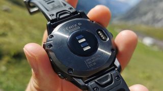 Baksidan av Garmin Fenix 6 som visar dess sensorer, med en somrig bergsmiljö i bakgrunden.