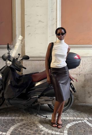 a photo of a woman wearing minimalist mule sandals with a brown leather skirt and white turtleneck