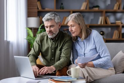 Mature couple on sofa in cozy living room using laptop and papers to review bills, budget and retirement plans, focused on savings, mortgage and investments for future