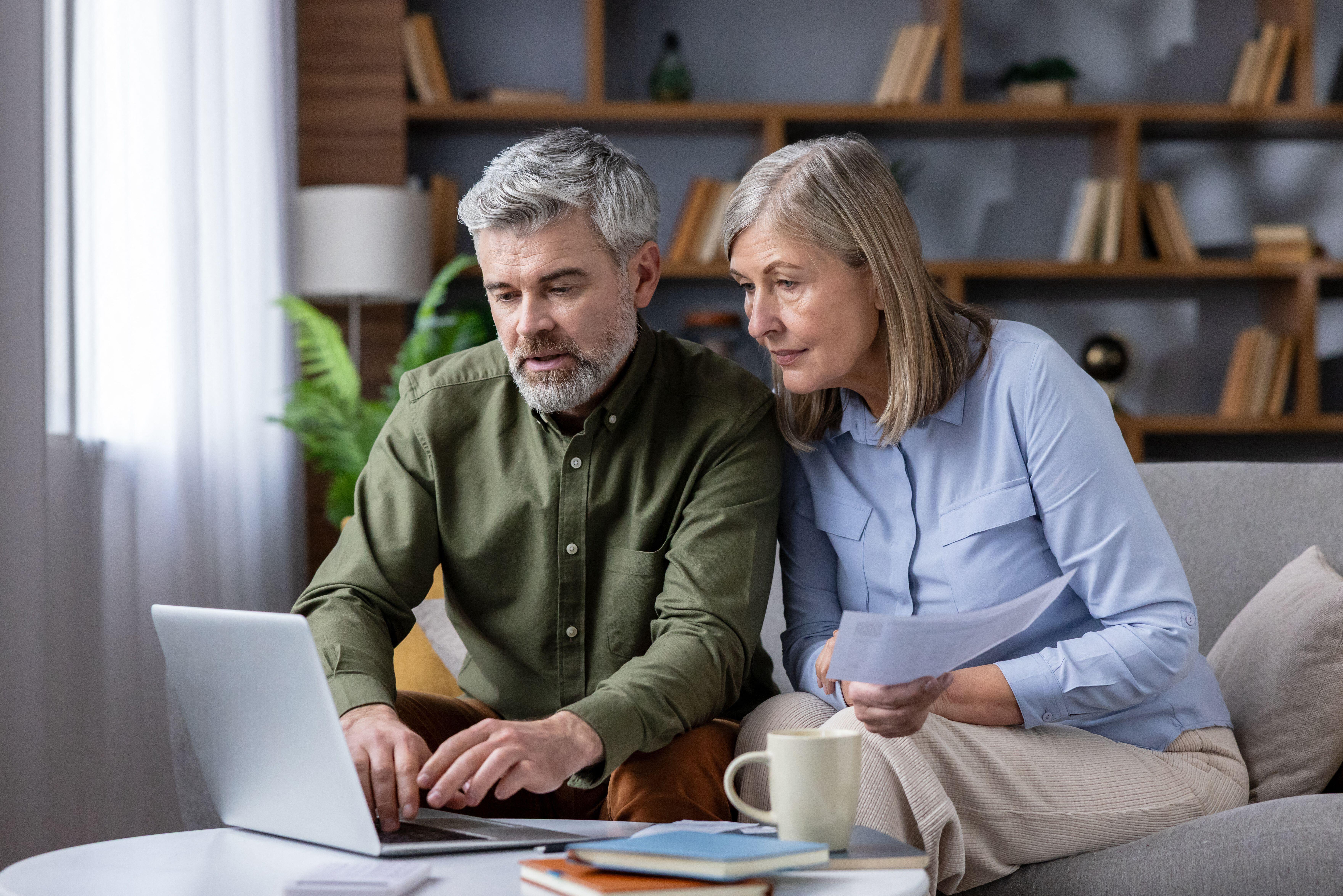 Mature couple on sofa in cozy living room using laptop and papers to review bills, budget and retirement plans, focused on savings, mortgage and investments for future