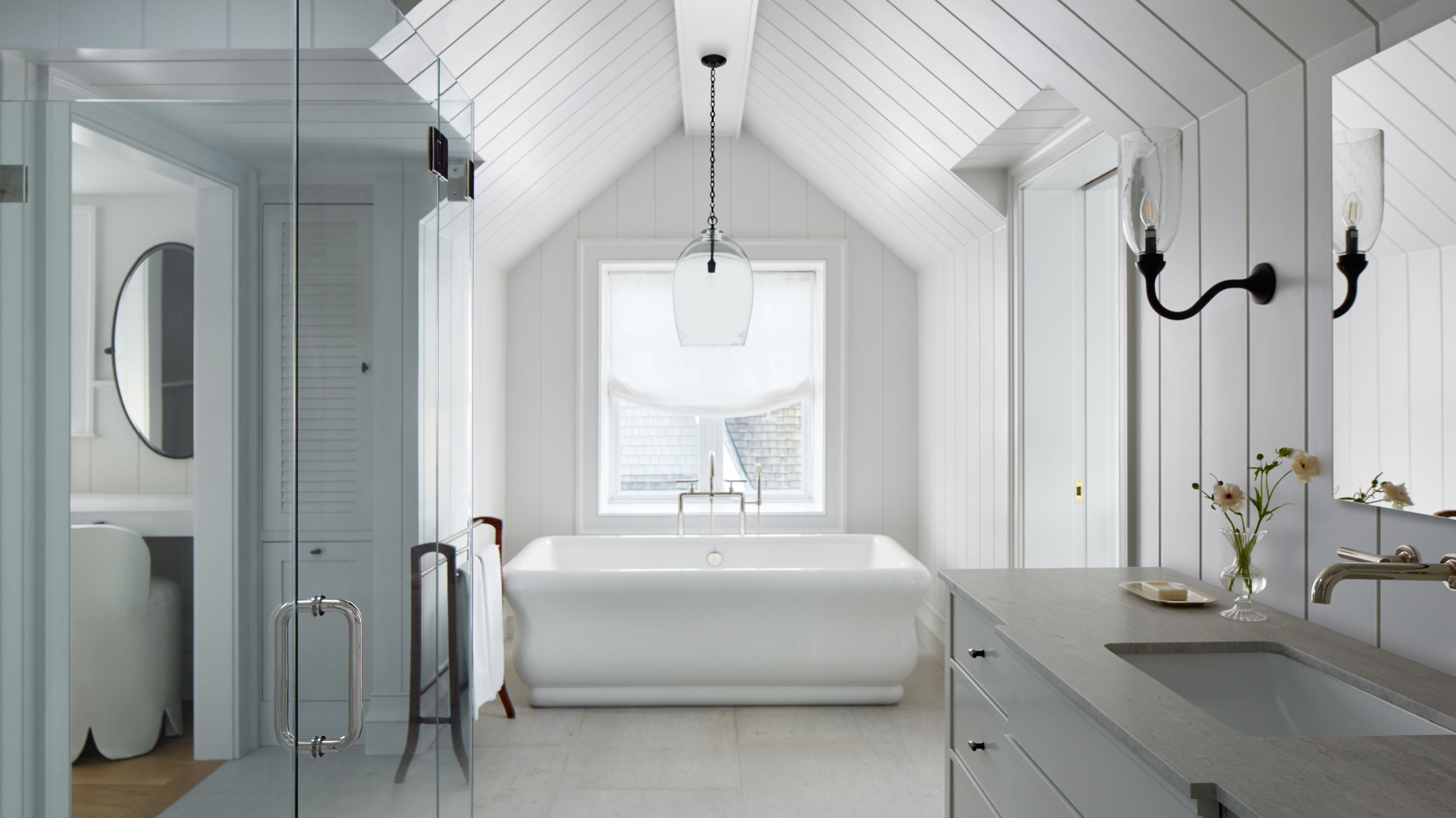 A bright, symmetrical bathroom featuring a white soaking tub centered under a vaulted shiplap ceiling. A glass pendant light hangs above the tub, flanked by a marble vanity and a glass-enclosed shower.
