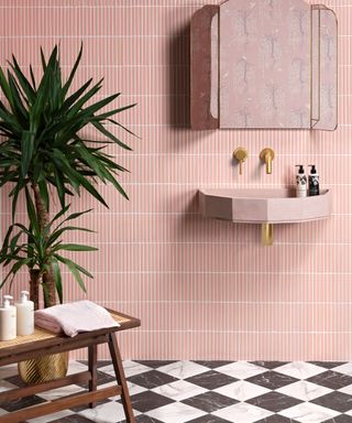 A bathroom with light pink tiles on the wall, a wall mirror, a pink sink with soap on it, and a marble checkerboard floor with a wooden stool and a tall spiked green potted plant on it