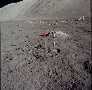 A red/orange flag stands on the lunar surface. The lunar roving vehicle is visible in the distance.