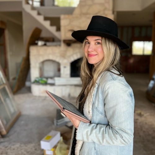 Image of a woman in a light denim jacket and a black cowboy hat standing in a home that is under construction with an iPad in her hand.