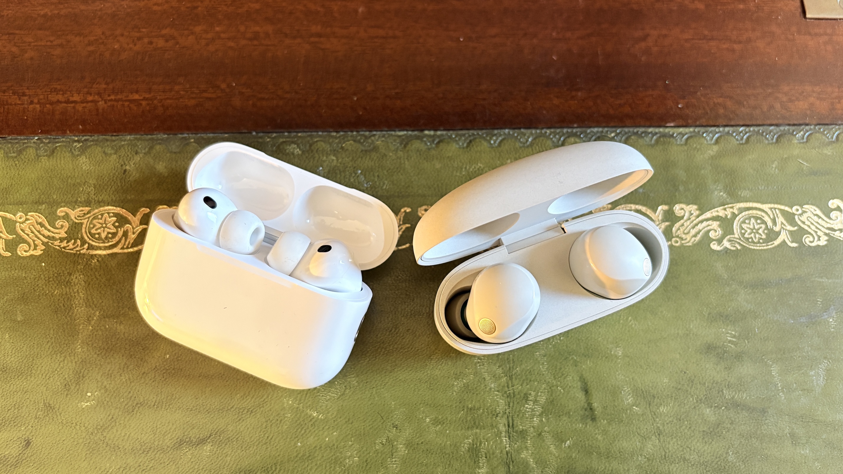 AirPods Pro 3 and Sony WF-1000XM5 earbuds in their open charging cases side by side on a green surface.