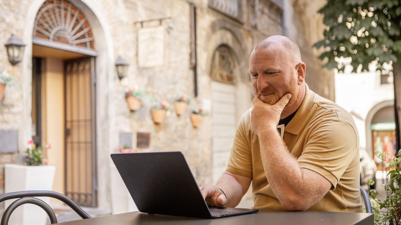 An older man works on a laptop while sitting outside an Italian villa.