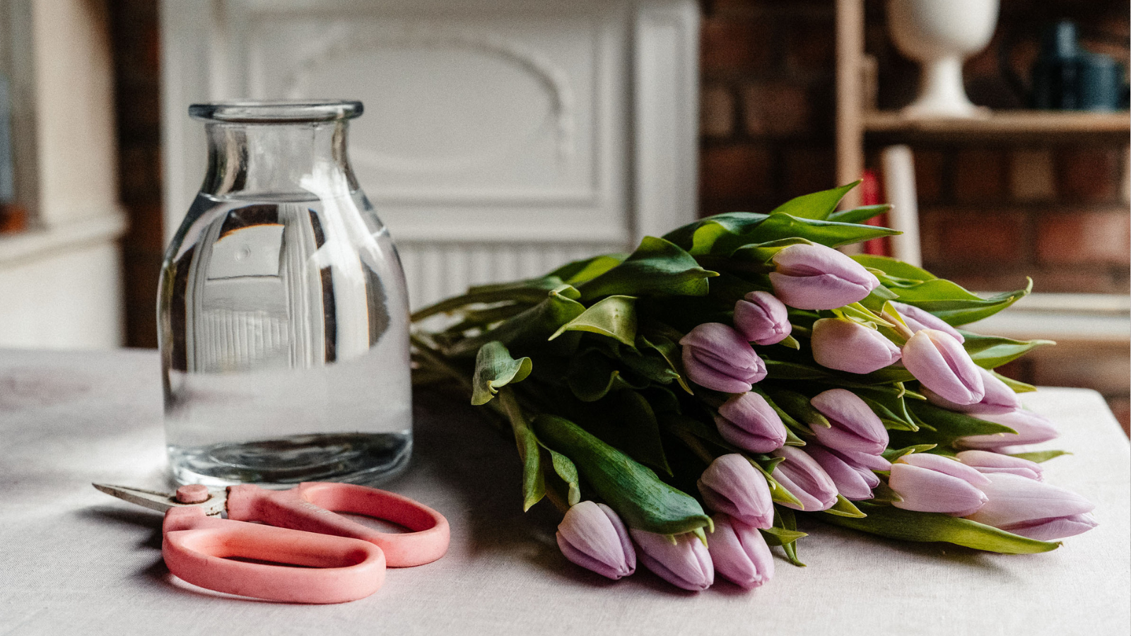 Lilac tulips on blush tablecloth with pink scissors and vase of water