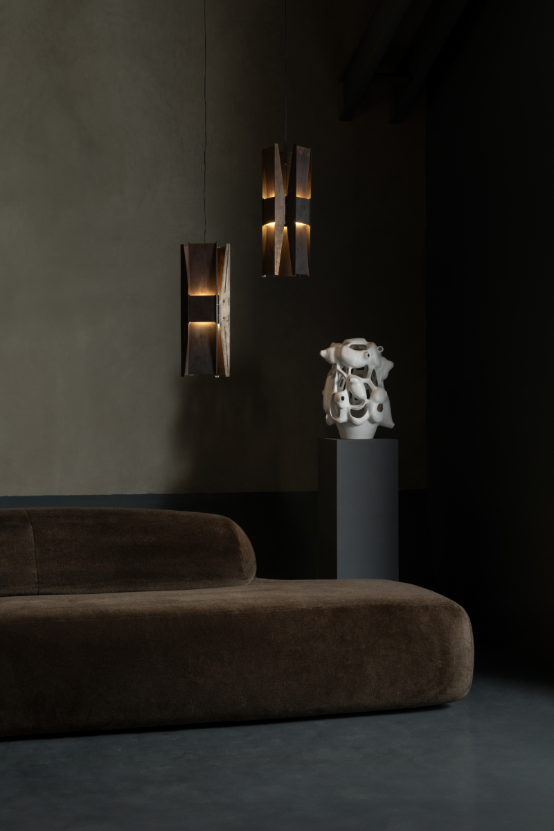 A dimly lit design showroom boasts a sculptural brown velvet sofa with an oversized shape, pendant lighting matching its color, and a podium topped by a whimsical white sculpture.