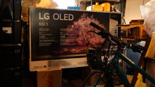 A box for the LG C1 OLED TV in a basement. It is sitting atop two cardboard boxes and in the foreground is a blue bicycle. The TV box is surrounded by other storage items.