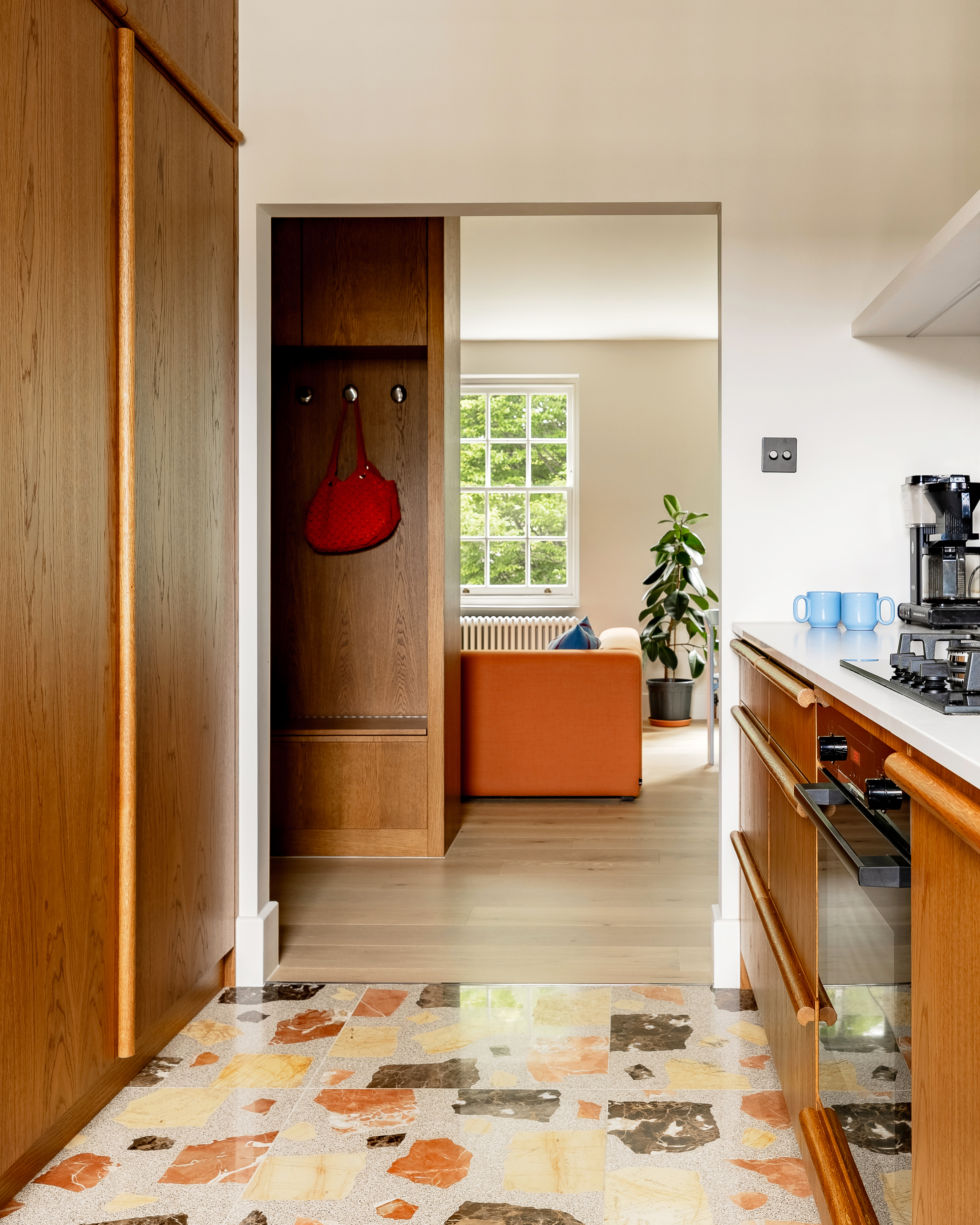 a kitchen with a large terrazzo floor, wood doors with dowel handles, and a view through to the living room with an orange sofa