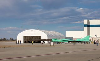 The profile of a jet with pale green panel coverings with sleek, sharp edges and angles, and a long pointed black nose, is rolled outside a hanger. Personnel walk scattered about the tarmac.