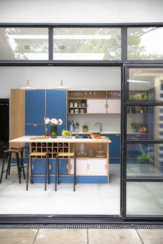 Shot from outside showing kitchen with blue and pink Formica plywood units, white worktop, industrial-style bar stools and open oak shelving, framed by metal Crittall-style doors