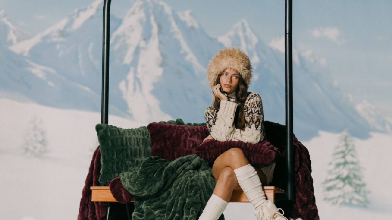 A woman sitting on a ski lift draped in a dark green and plum coloured blanket from Lola Blankets, in front of a wintery mountain backdrop.