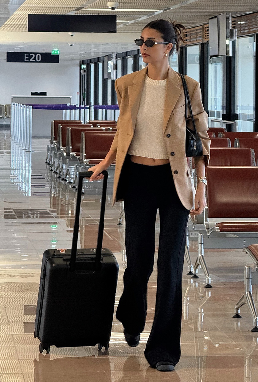 Summer airport outfit is shown in a street style photo of a French woman wearing black sunglasses, gold earrings, a white crewneck, black trousers, black flats, a black shoulder bag and a black carry on suitcase