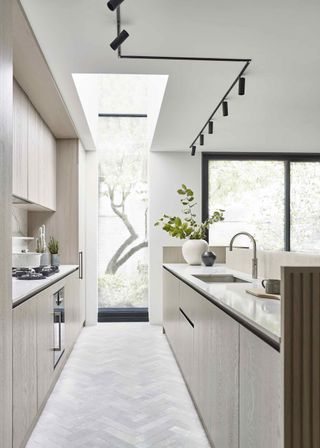 A galley layout and double height glazing. This small kitchen has a countertop with a sink on the right and on the left is a countertop with a gas stove. On the ceiling are spotlights.