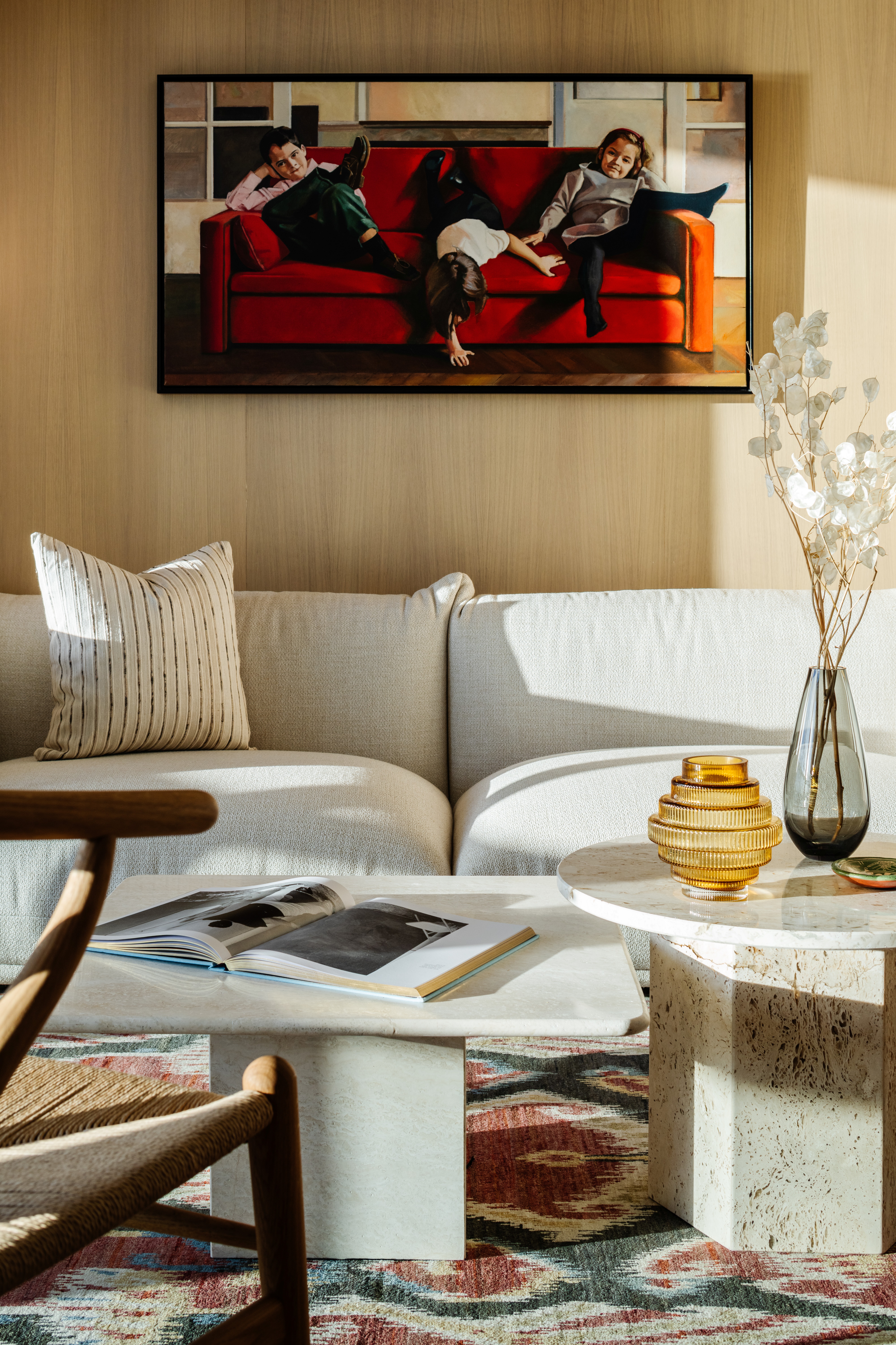 A naturally lit interior with a lounge sofa in a soft shape, wood-paneled walls, wood furniture along with marble accents, eccentric artwork, and a pendant paper lantern.