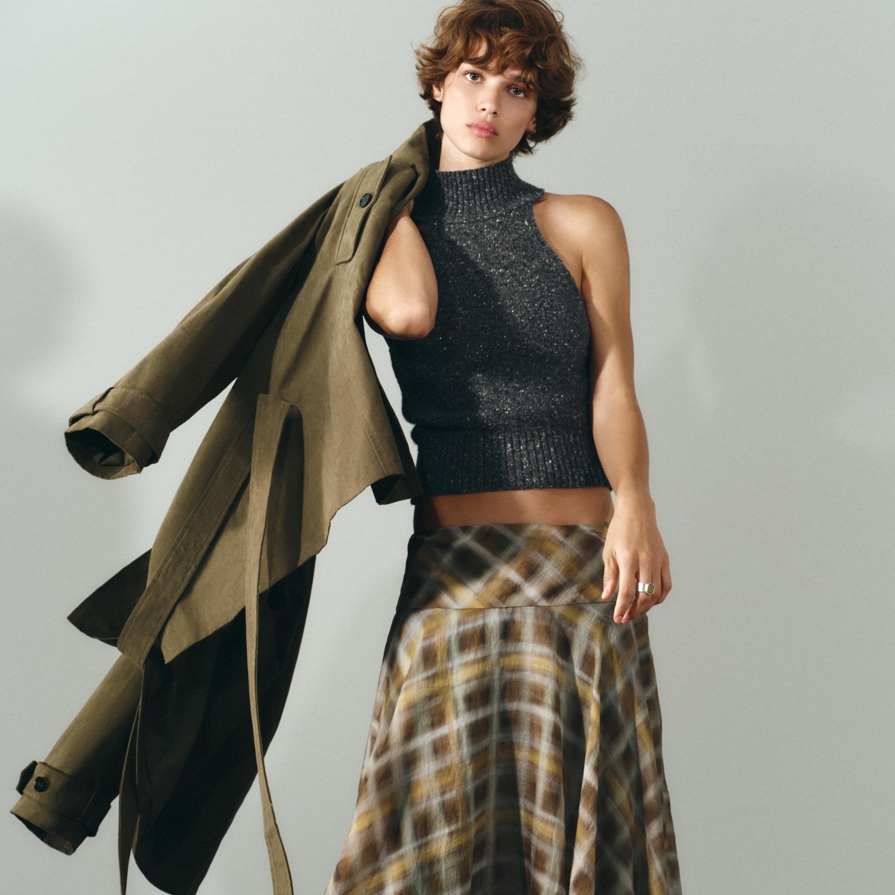 Model wearing a grey knitted tank top, checked skirt and holding a trench coat.