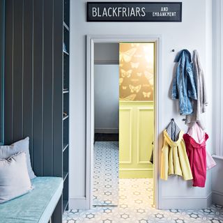 A boot room with dark grey pigeon holes, insect motif wallpaper decor, yellow wall panel decor and children's coats hung on pegs