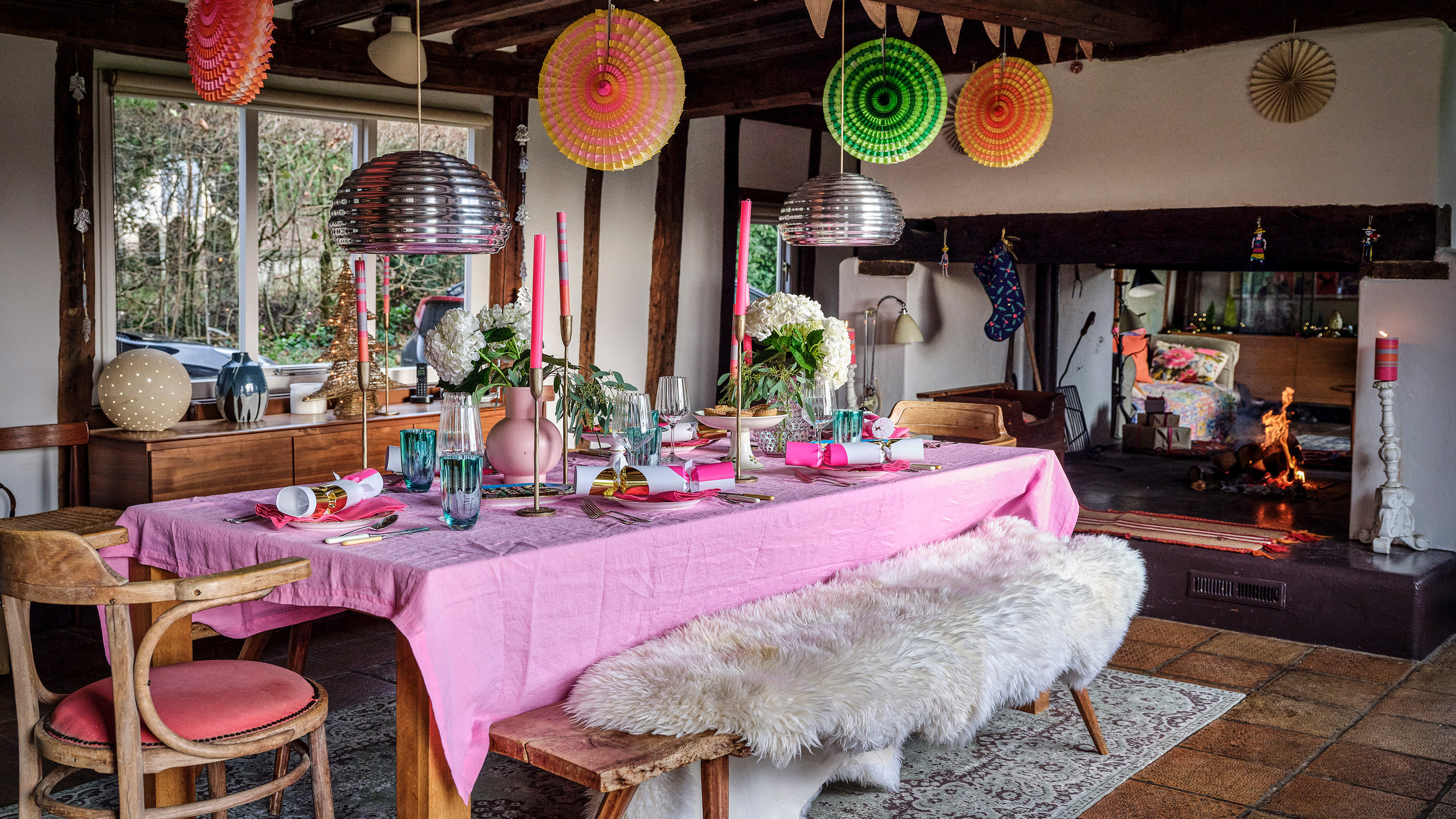 dining room in barn with long table with pink tablecloth bench seating with a fur cover and colourful paper decorations hanging from the ceiling 