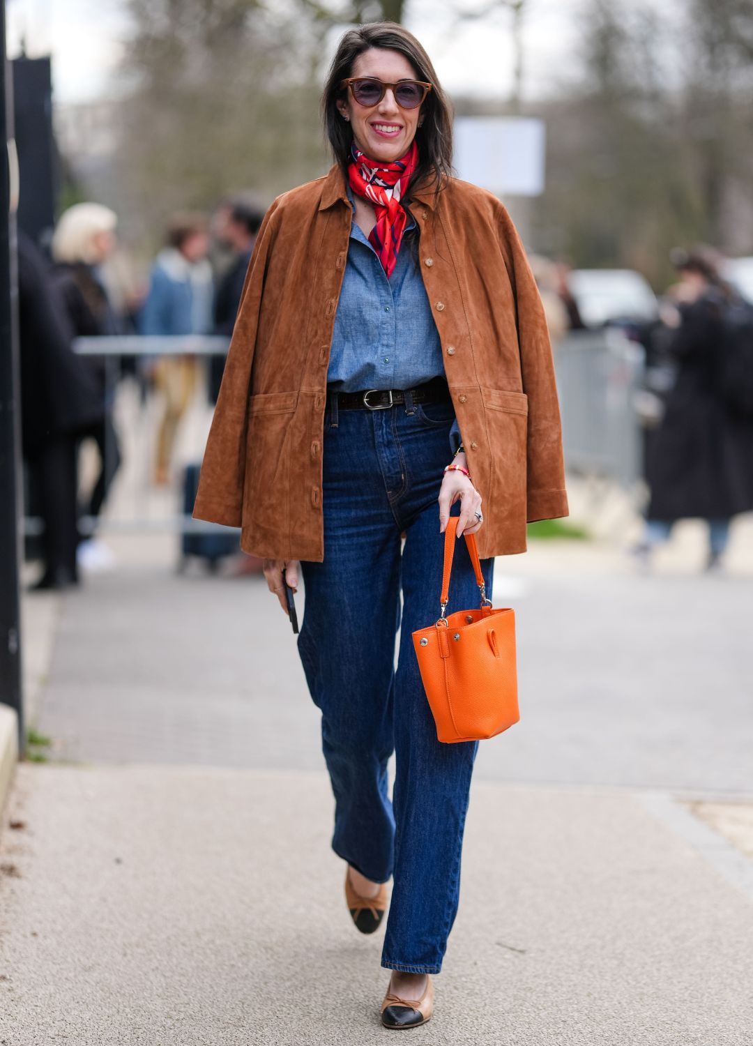A guest wears dark brown sunglasses, bright cherry red pattern/print scarf around neck, navy blue buttoned up long sleeve shirt, dark brown oversized suede jacket, navy blue loose denim jean pants, shiny black leather bag, bright orange leather bag, cream black ballerina flats Chanel leather shoes, outside Lacoste, during the Paris Fashion week Women&#039;s Fall/Winter 2025-2026 on March 9, 2025 in Paris, France
