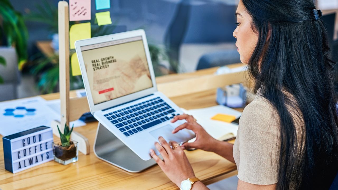 Woman with long black hair using the touchpad on a laptop raised on a laptop stand. A sign next to her reads &quot;good coffee always&quot;