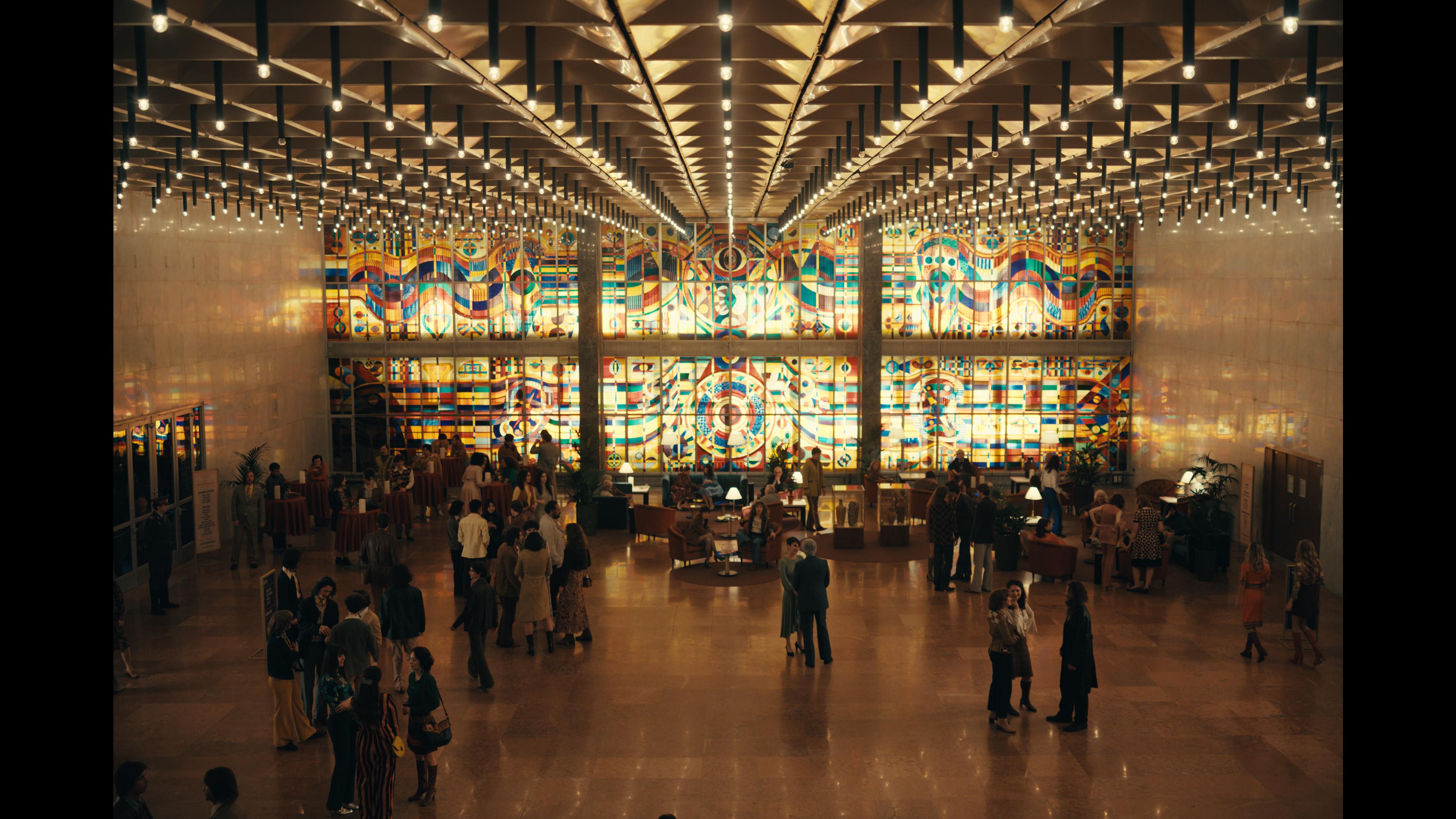 A series of film stills and photographs from a 1970s-inspired set featuring vibrantly decorated interiors with layered furniture and warm lighting, along with characters dressed in vintage clothing whether indoors or at parks, in cards, or at markets outdoors.