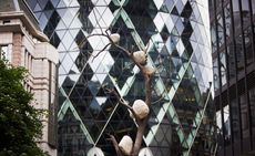 A tree-like sculpture with rocks placed on different levels of the tree branches. Photographed during the day with a tall glass reflective office building in the background