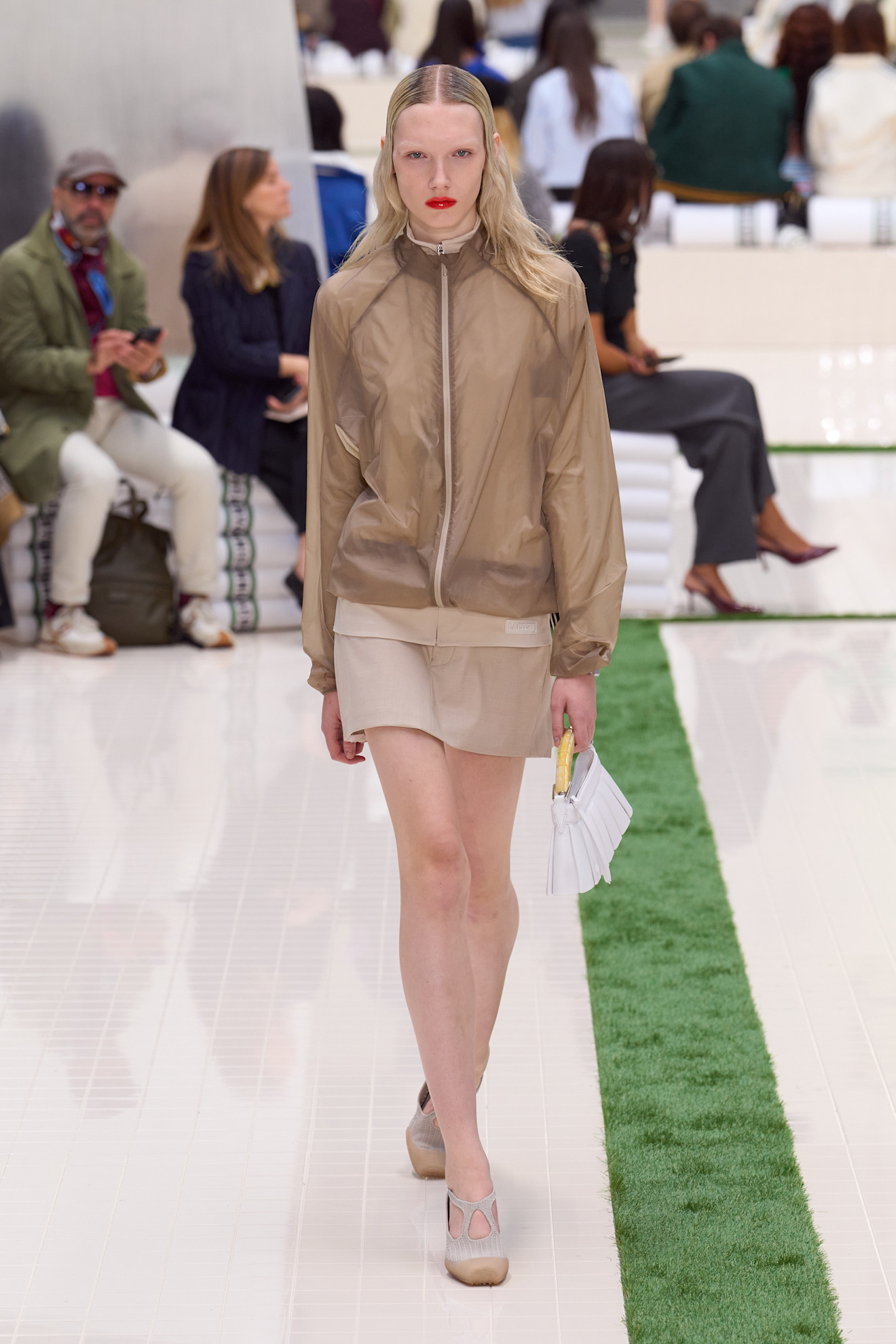 Layering staples for spring 2026 is shown in a photo from Lacoste's runway show, with a model walking while wearing a light tan bomber jacket layered over a tan turtleneck sweater, worn with a tan mini skirt, tan pumps, and a white handbag
