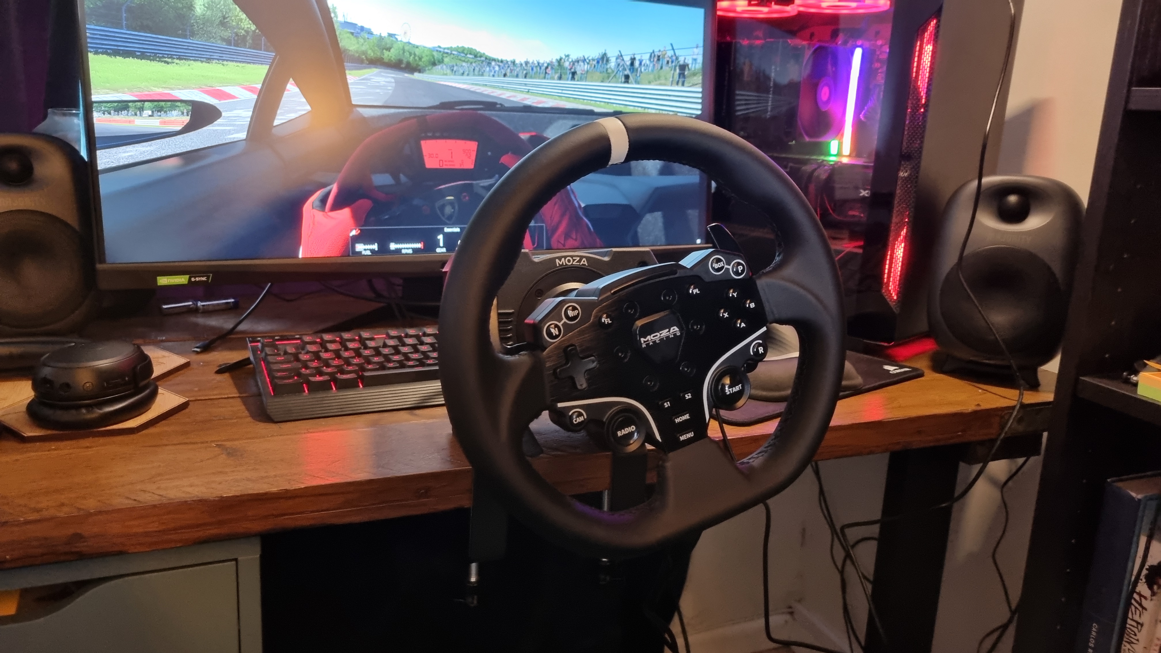 The MOZA R5 wheelbase mounted to a wooden desk with desk clamps, with a monitor and gaming rig showing a Lamborghini in Assetto Corsa at the Nürburgring