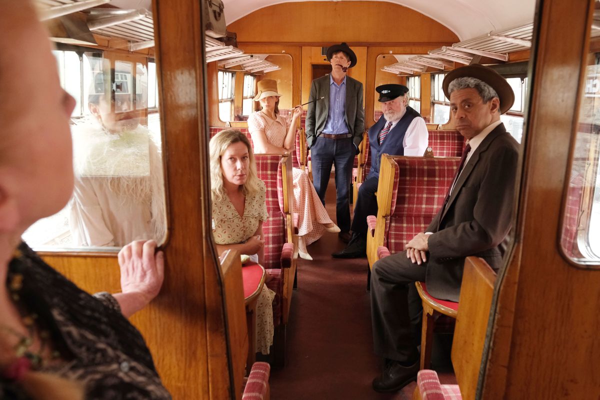 Beyond Paradise season 2 episode 1: an interior shot of a vintage train carriage with DI Humphrey Goodman (Kris Marshall) standing in the background, wearing a trilby and with a pipe hanging out of his mouth. Various actors in approximately 1930s-era attire are sitting in the carriage, and Margo Martins (Felicity Montagu), in modern clothing, is just visible in the foreground, leaning against the dividing panel