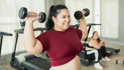 A woman stands in a gym with dumbbells held at shoulder height. Her elbows are pointing down, out to the sides, and she is smiling. Behind her is a row of treadmills, and a woman sitting down on one of them.