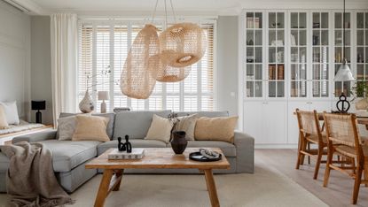 Living room with grey walls, white shutters and curtains, white glazed cabinets and grey corner sofa with wood coffee table and rattan pendant lights overhead