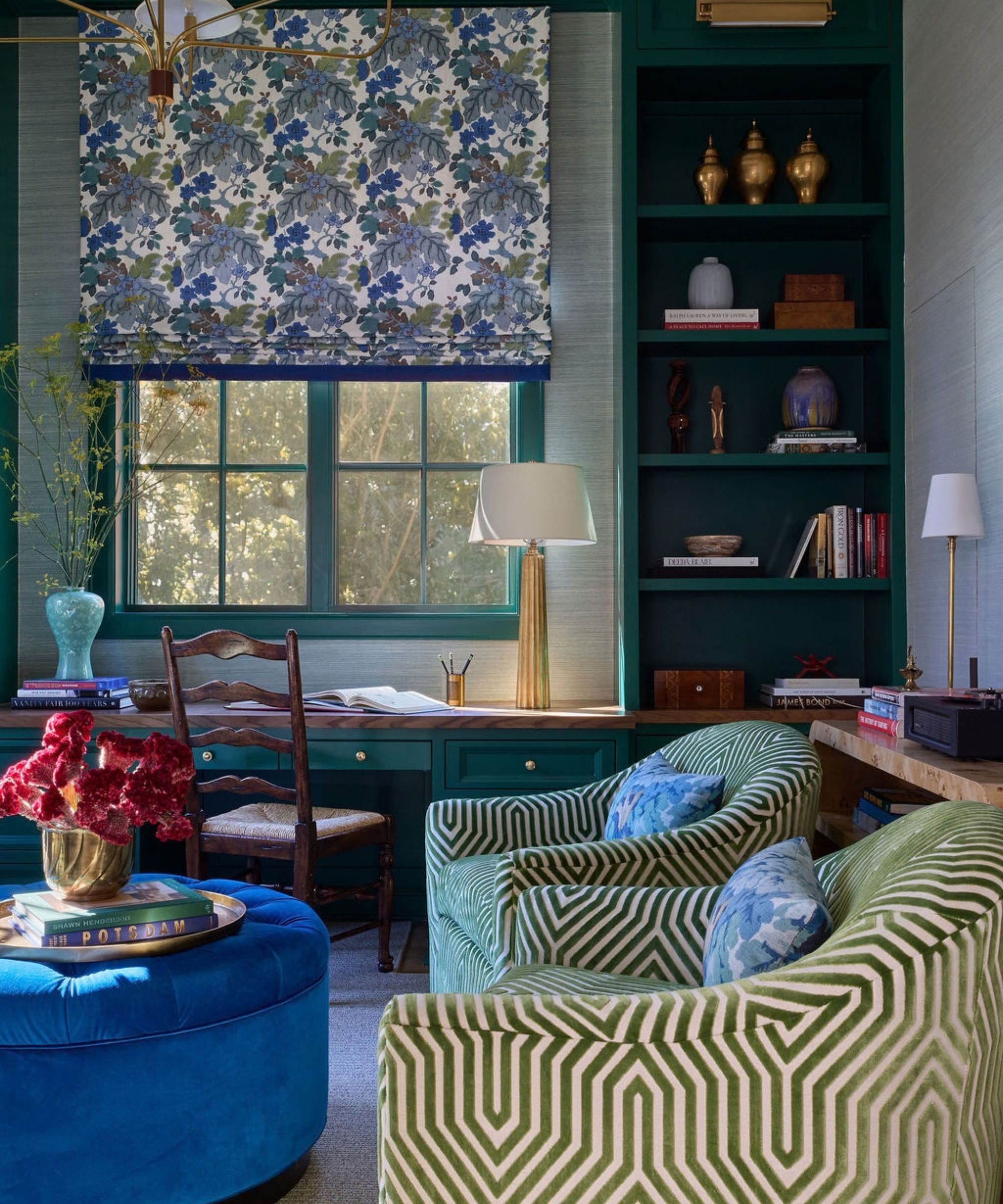 Dark green study featuring built-in bookshelves, geometric pattern swivel chairs, a blue velvet ottoman, and a patterned roman shade