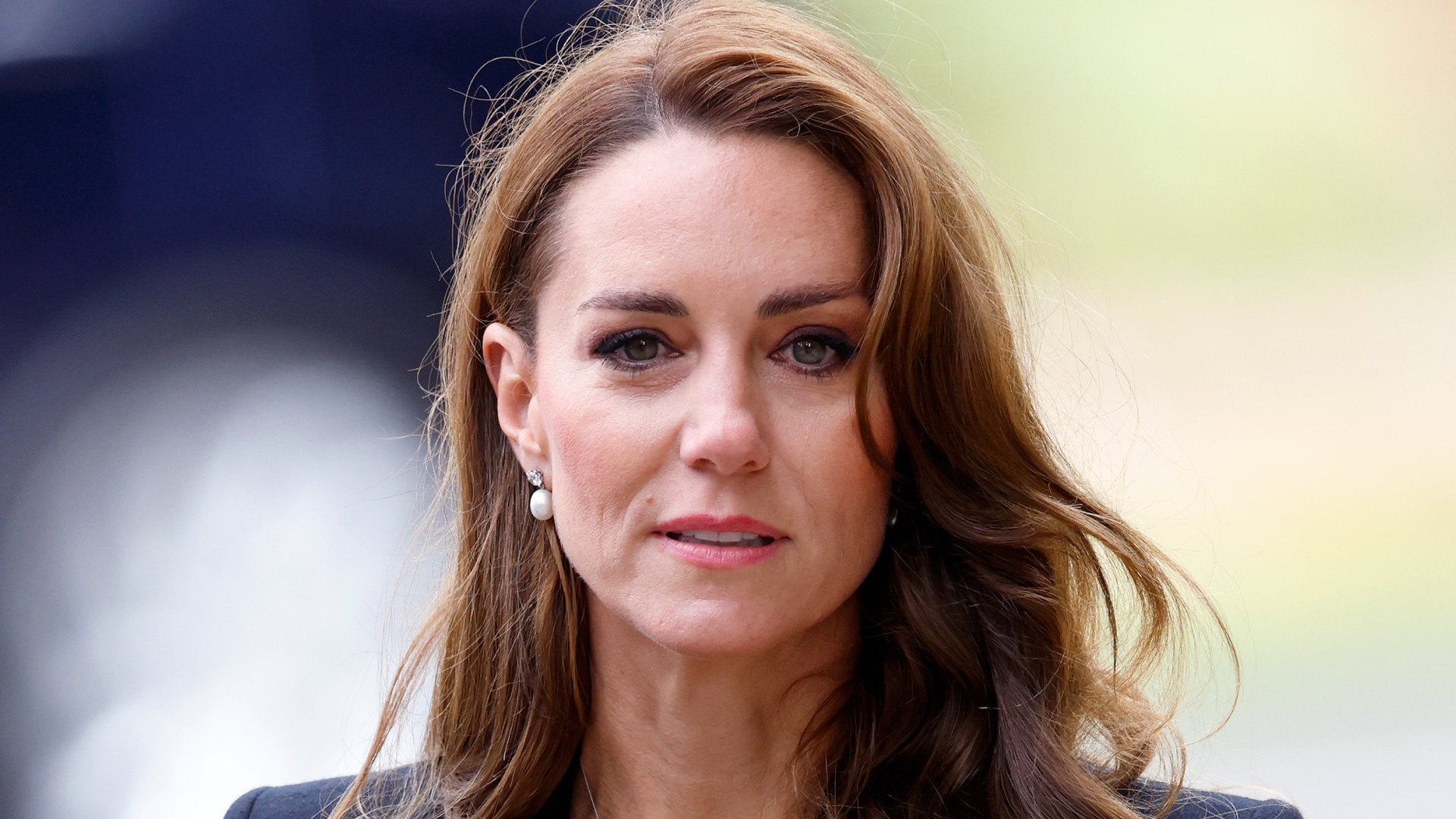 Princess Kate Middleton wears a black blazer and black top and has long wavy brunette hair featuring blonde highlights