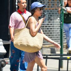 Zoe kravitz wears a blue baseball cap, lavendar dress, suede sandals, and a raffia tote bag.