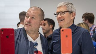 Two businessmen, Jony Ive and Tim Cook, talk with out of focus iPhones on stands in the foreground