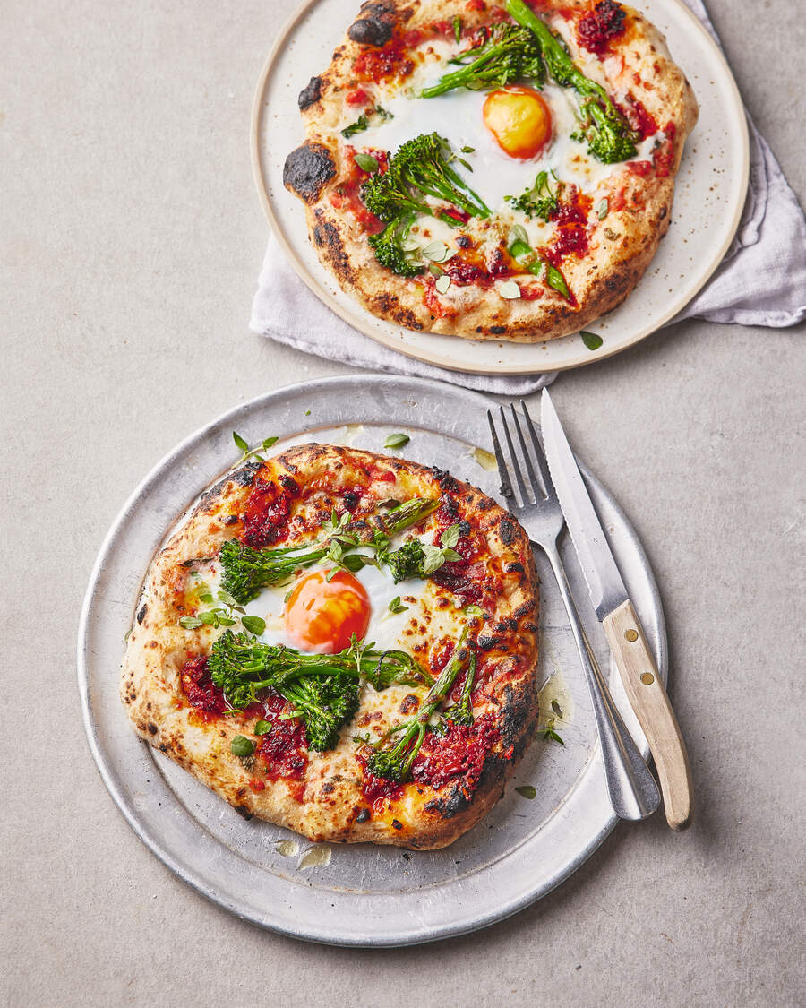 Two pizzas topped with spicy salami paste, an oozy egg, and broccolini, served on plates, perfect for brunch or dinner.